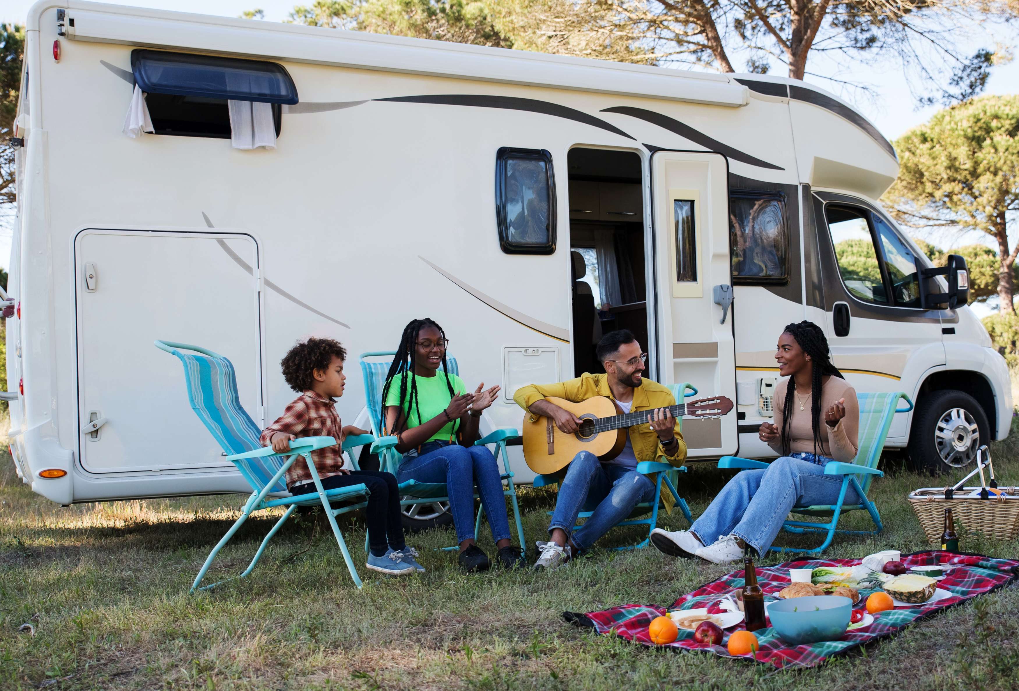 Four friends enjoying a musical gathering outside a camper van, surrounded by snacks and nature.