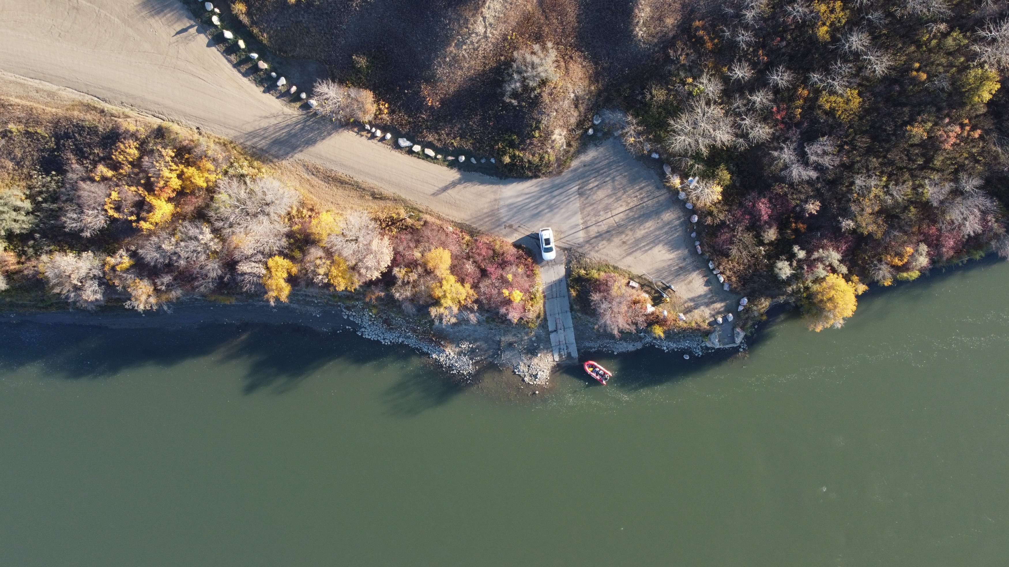 Aerial view of a riverbank with autumn foliage, a vehicle, and a small boat near the water.