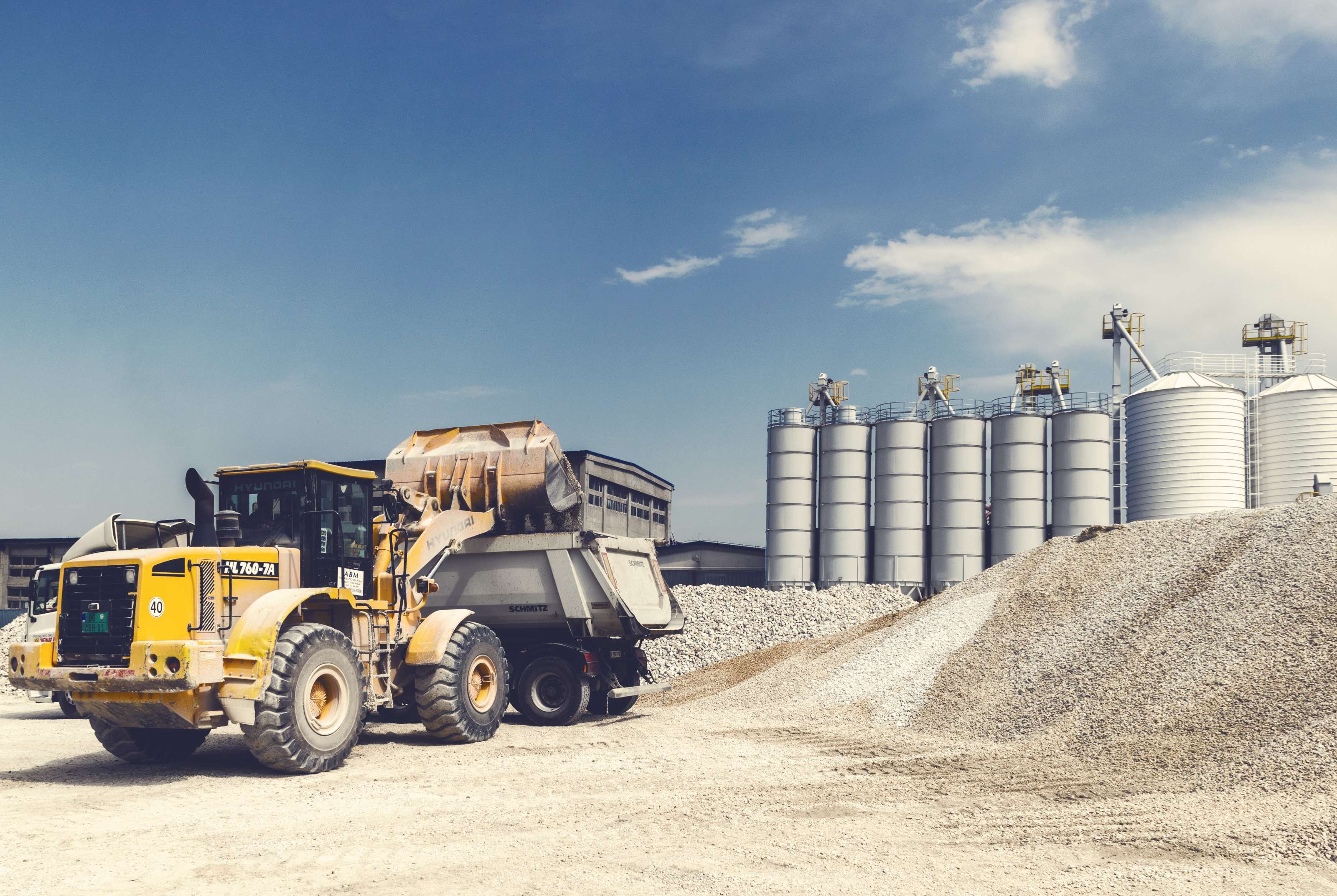 A loader fills a dump truck with gravel, surrounded by piles on the left and metal silos in the background.