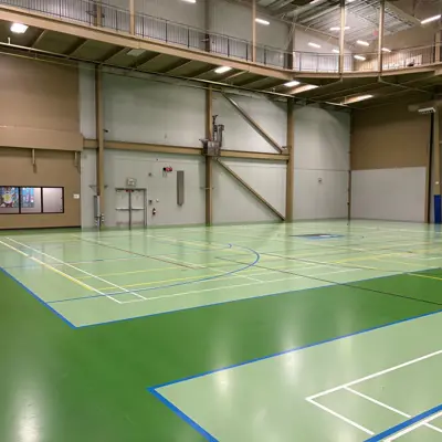 Empty indoor gymnasium with green multi-sport courts, overhead lighting, and a scoreboard.