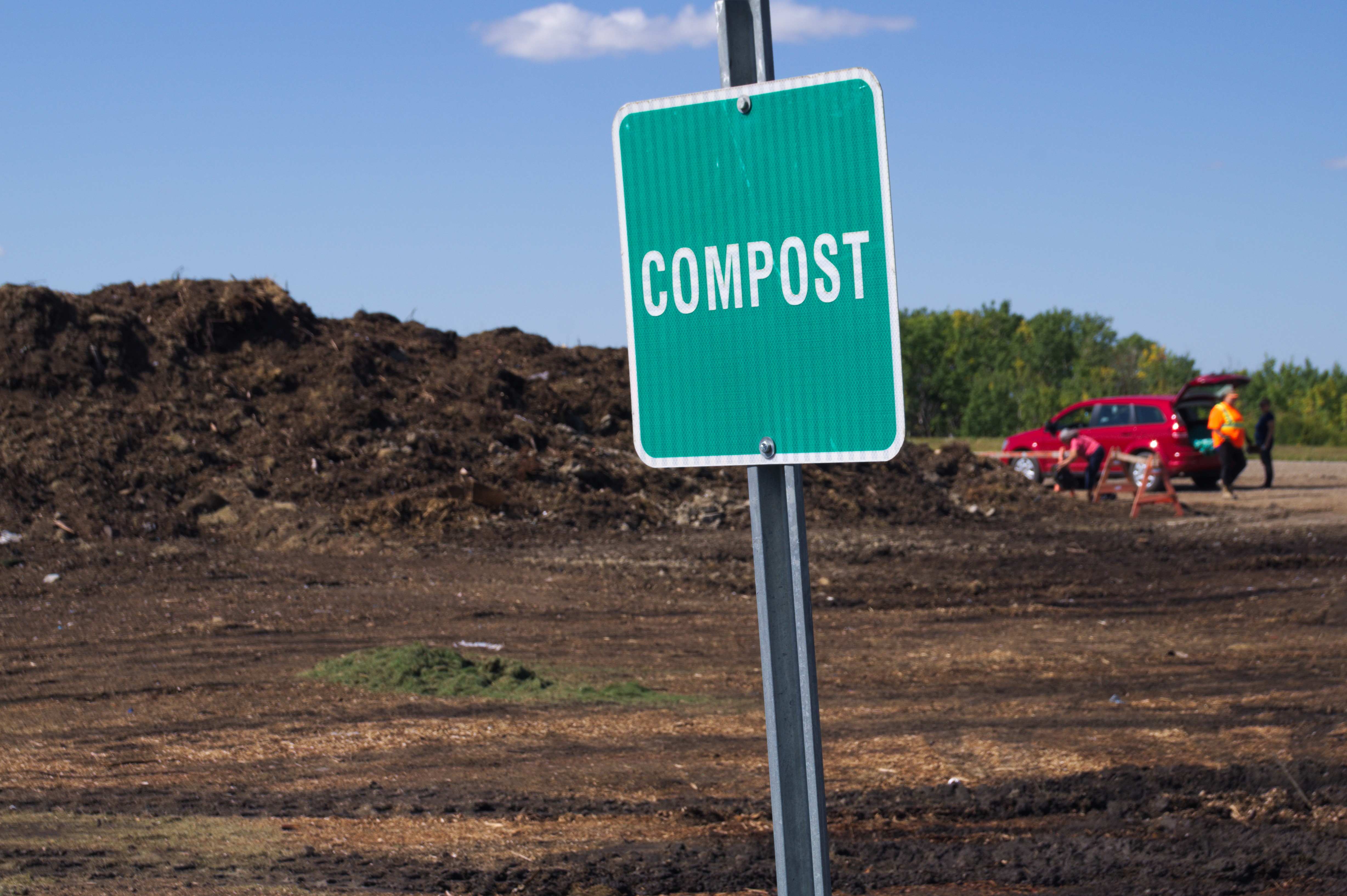 A green sign in the foreground that says Compost, while in the background is a large compost pile and a person in a red vehcile next to it adding to the pile.