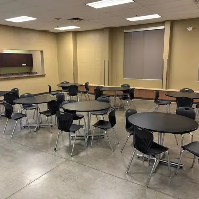 Cafeteria room with round tables, black chairs, serving window, and concrete floor.